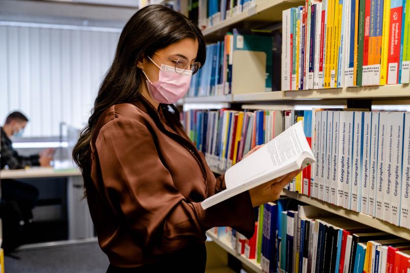 Studentin mit Mund-Nase-Schutz in der Bibliothek, im Hintergrund sitzt ein Studierender mit Maske an einem Tisch.