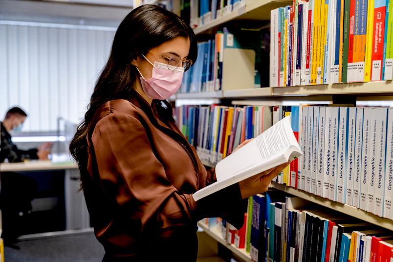 Studentin mit Mund-Nase-Schutz in der Bibliothek, im Hintergrund sitzt ein Studierender mit Maske an einem Tisch.