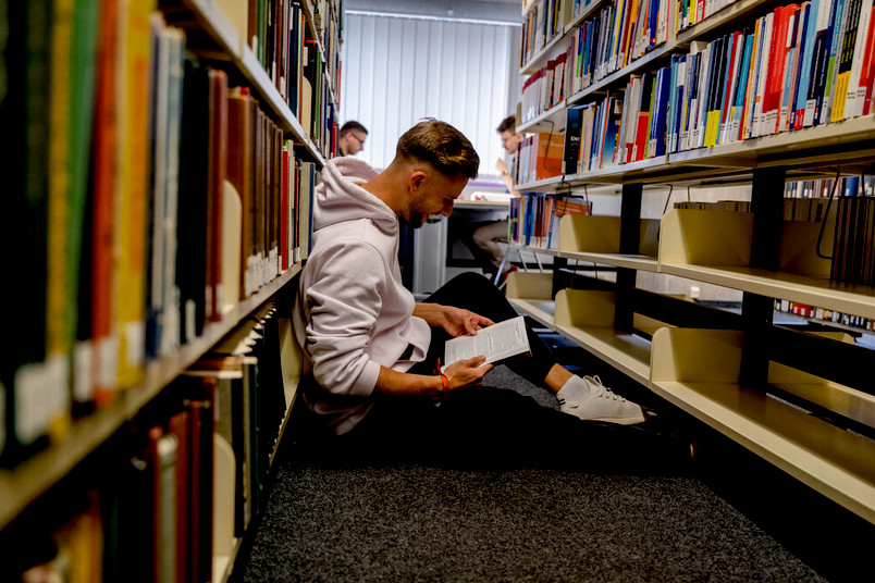 Student sitzt auf dem Boden der Bibliothek zwischen zwei Bücherregalen und liest