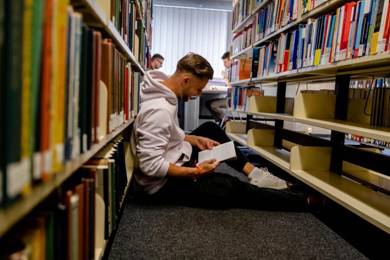 Student sitzt auf dem Boden der Bibliothek zwischen zwei Bücherregalen und liest