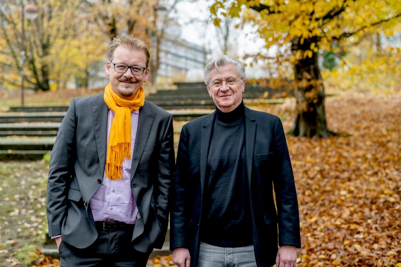 Klaus Oschema steht im grauen Anzug mit lilanem Hemd und orangem Schal lächelnd auf dem Campus der Ruhr-Universität Bochum. Neben ihm steht Bernd Bastert im schwarzen Rollkragenpullover mit grauem Jackett und guckt freundlich in die Camera.