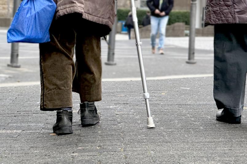 Eine ältere Person läuft mit einem Gehstock über die Straße. Sie ist von hinten zu sehen.