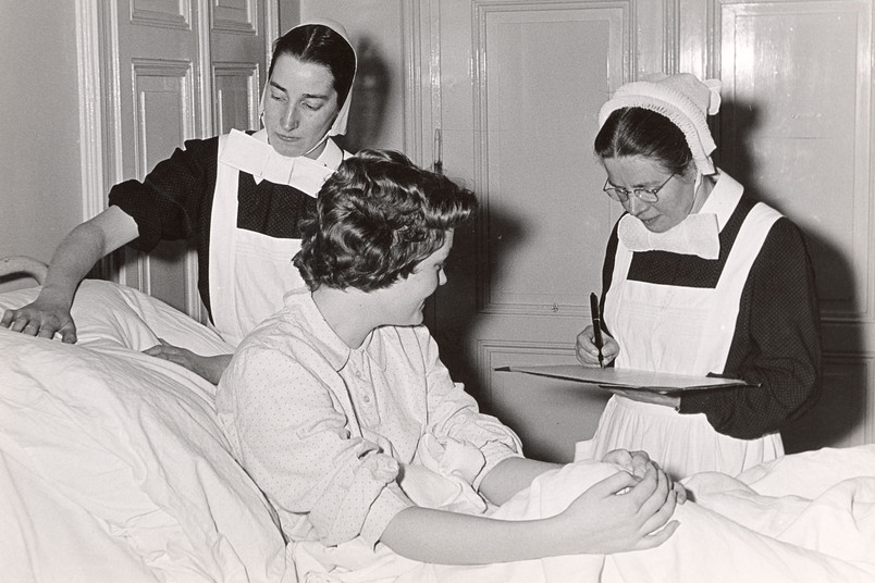 Schwarzweiß-Foto von zwei Schwestern in Tracht mit Haube vor einer Person im Krankenbett.