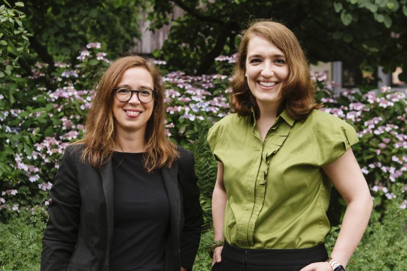 Annett Schmeck und Nora Čavara stehen nebeneinander vor grünem Hintergrund und lachen in die Kamera.