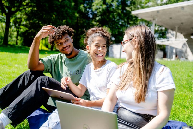 Drei Personen sitzen auf deiner Wiesen und lächeln, zwei Personen haben ein Tablet oder Laptop auf dem Schoß.