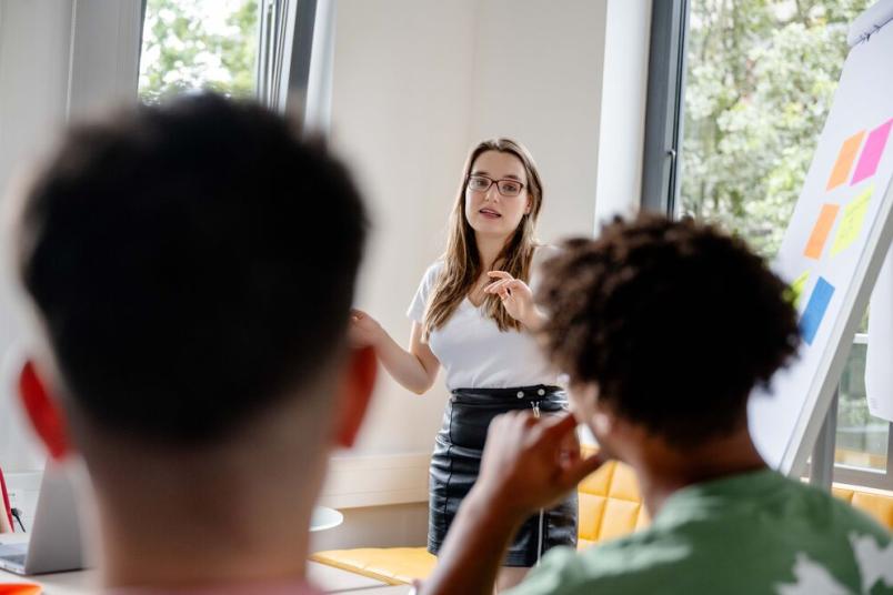 Lehrende steht an einem Flipchart und erklärt Studierenden etwas.