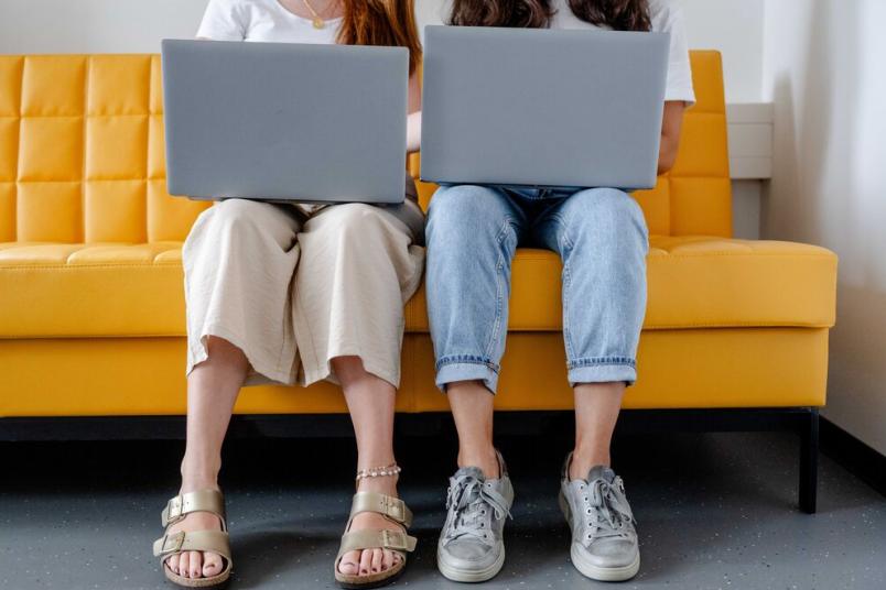 Studentinnen sitzen nebeneinander mit Laptops auf dem Schoß. 