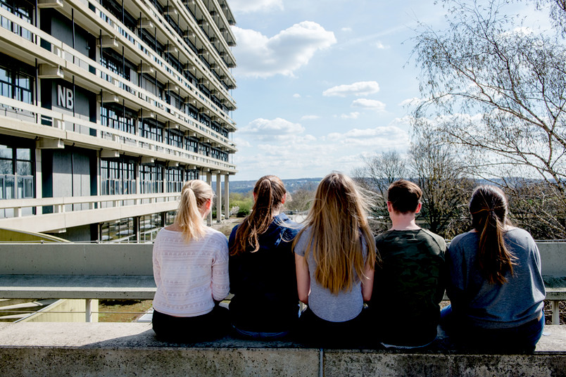 Fünf Personen sitzen mit dem Rücken zur Camera an einem Gebäude der RUB und Blicken in das Tal dahinter.