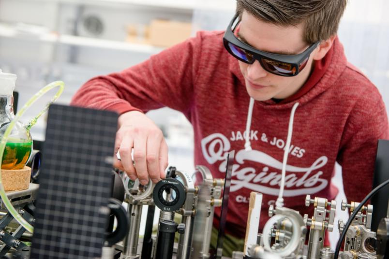 Junger Mann mit Schutzbrille beugt sich über ein Experiment an einem Laser.