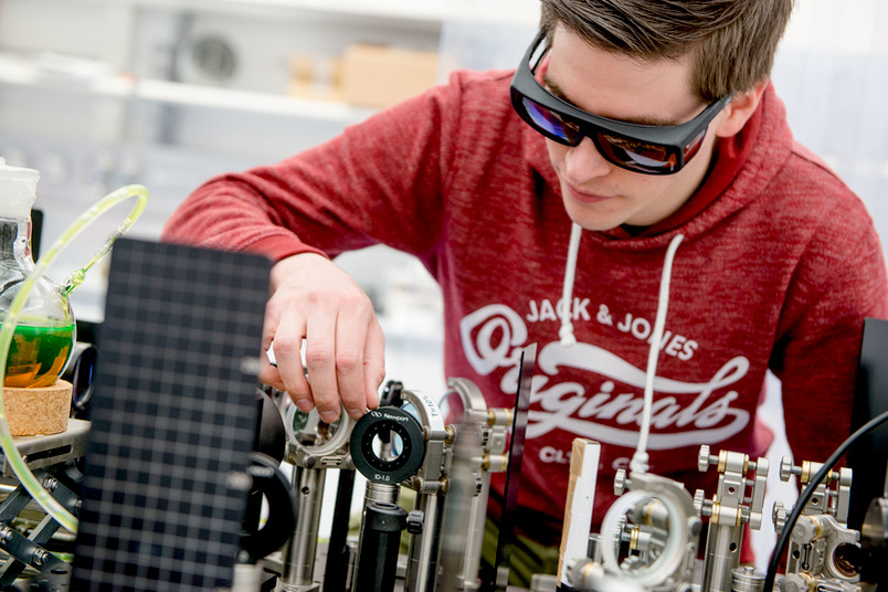 Junger Mann mit Schutzbrille beugt sich über ein Experiment an einem Laser.