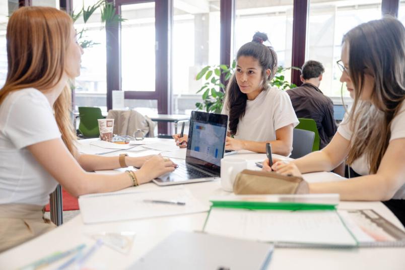 Drei Studentinnen lernen zusammen. Ein Laptop steht auf dem Tisch. Bücher liegen daneben. 