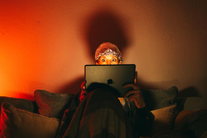 Das Bild zeigt einen Kopf von einer Frau, die einen Alu-Hut trägt und mit weit aufgerissenen Augen auf einen Bildschirm eines Laptops starrt.