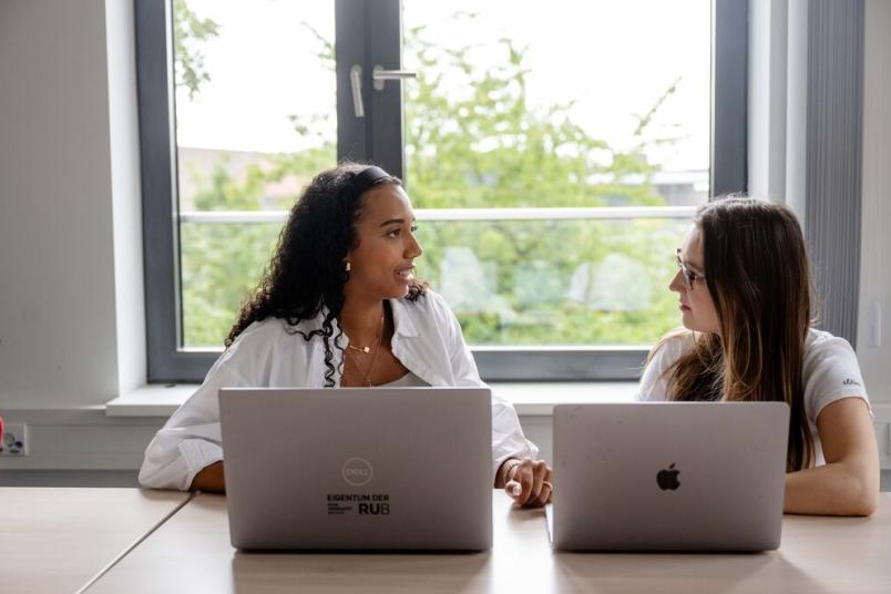 Zwei Frauen sitzt an zwei Laptops nebeneinander und unterhalten sich. 