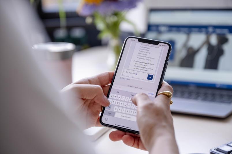 Blick über die Schulter einer Person. Sie hält ein Smartphone in der Hand. Darauf ist ein Chat mit dem LLM Mistral zu sehen.