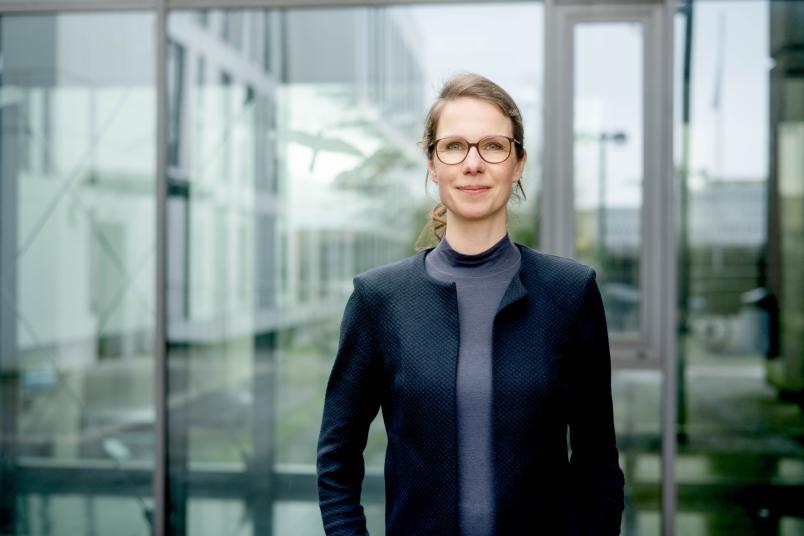 Frau mit dunklem Blazer, Brille und blondem Haar im Zopf