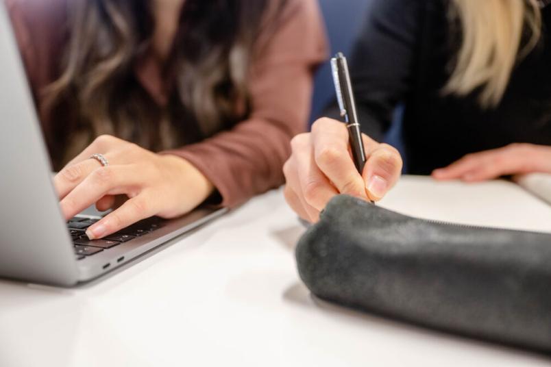Hände zu sehen. Eine schreibt auf einer Laptoptastatur. Eine schreibt mit einem Stift auf Papier. 