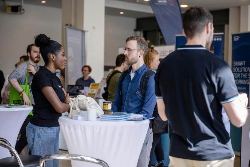 Ein Stehtisch an dem eine junge Frau und ein junger Mann stehen. Im Hintergrund sieht man Messetische und Aufsteller.