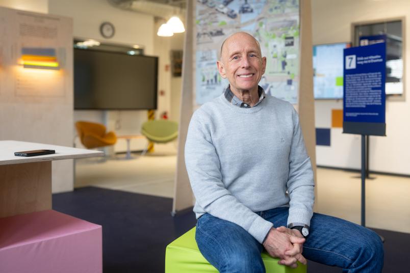 Mann im hellblauen Pulli auf einem hellgrünen Hocker vor einem Poster