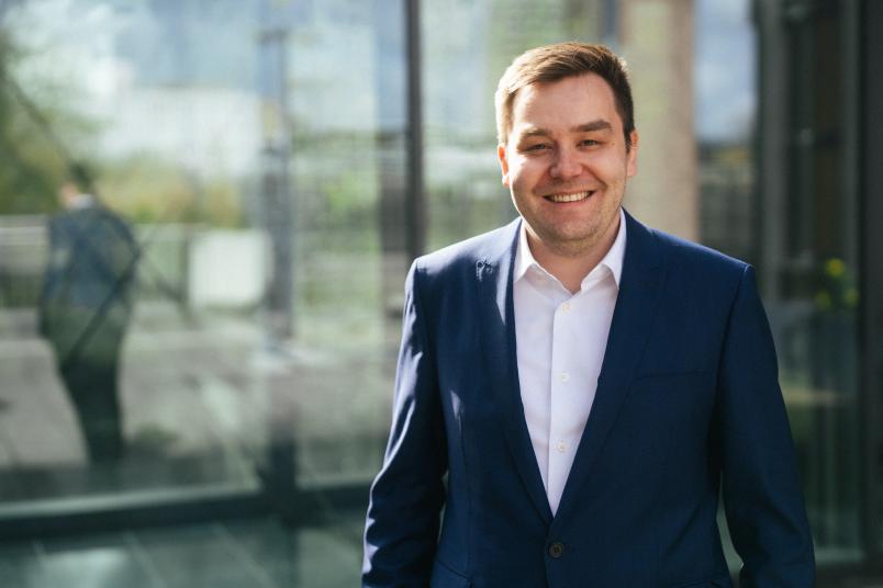 Ein junger, lachender Mann im weißen Hemd mit blauem Jackt vor einer spiegelnden Scheibe