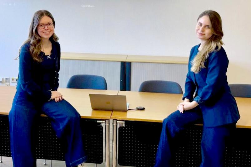 Zwei Studentinnen sitzen sich gegenüber auf Tischen. Beide sind in dunkelblau gekleidet.