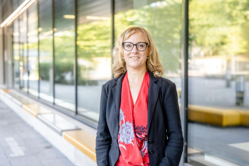 Blonde Frau mit schwarzem Blazer über roter Bluse steht vor Glasfenster.