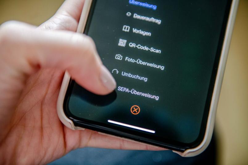 Nahaufnahme einer Hand, die ein Smartphone hält. Mit dem Daumen tippt sie auf den Menüpunkt "Überweisung".