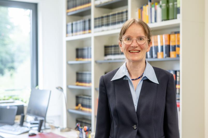 Eine Frau mit dunkelgrauem Jackett steht vor einer Bücherwand