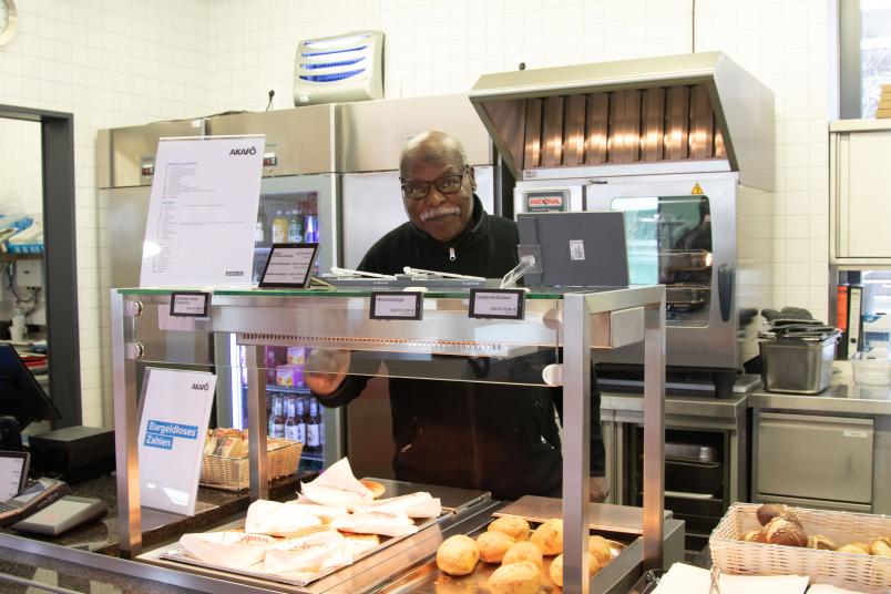Belgacem Derbal steht hinter dem Tresen der Cafeteria und blickt freundlich in die Kamera.