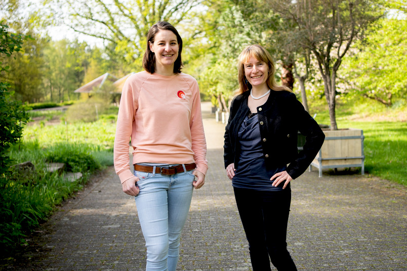 Judith Golda wears a pink pullover with blue jeans and ist standing next to Claire Douat between trees in the Botanic Garden in Bochum. Claire Douat wears a blue blouse with a black jacket and black jeans. They are smiling into the camera.