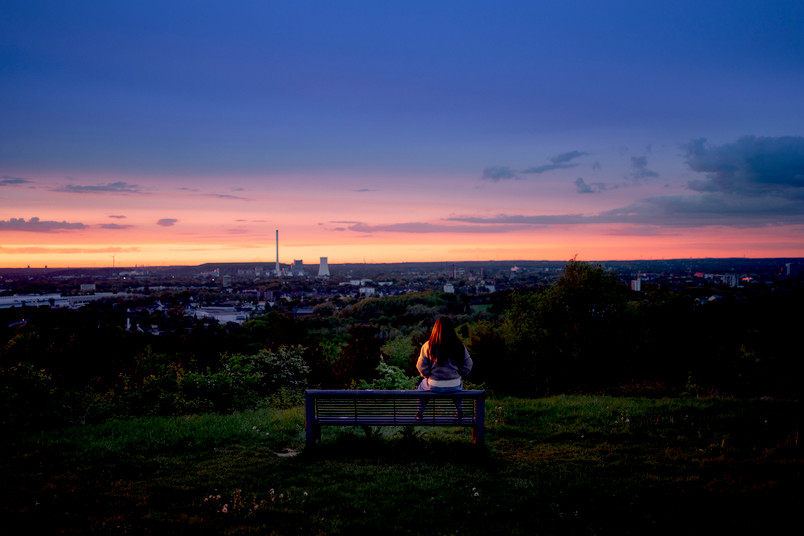 Eine einzelne Person sitzt auf einer Anhöhe auf einer Bank und schaut über das Ruhrgebiet.