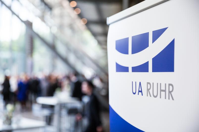 Das Logo der Universitätsallianz Ruhr in der Jahrhunderthalle. Im Hintergrund sind verschwommen Gäste zu sehen.