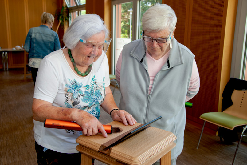 Zwei ältere Personen stehen vor einem einem Holztisch mit Tablet und halten einen Stab in der Hand