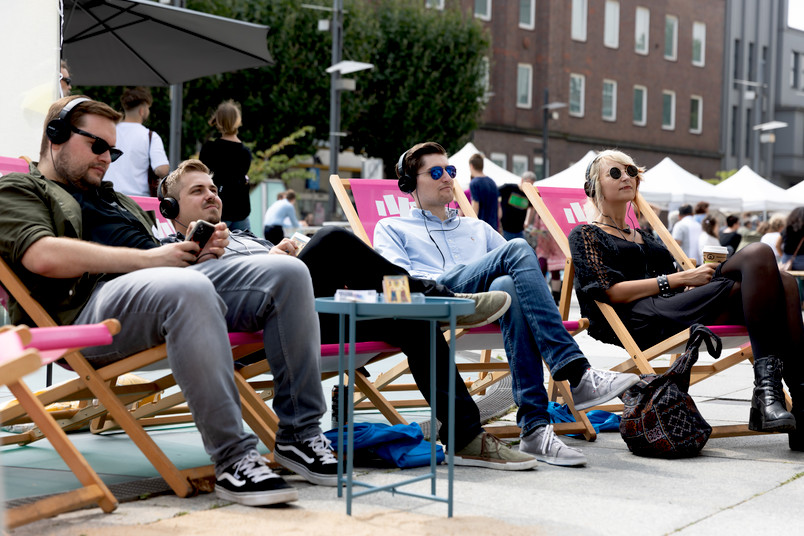 Junge Leute sitzen in pinken Liegestühlen auf dem Rathausvorplatz in Bochum und haben Kopfhörer auf.