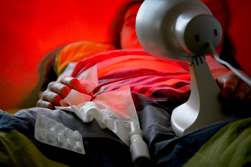 Eine Person liegt mit Rotlichtlampe und Taschentüchern im Bett