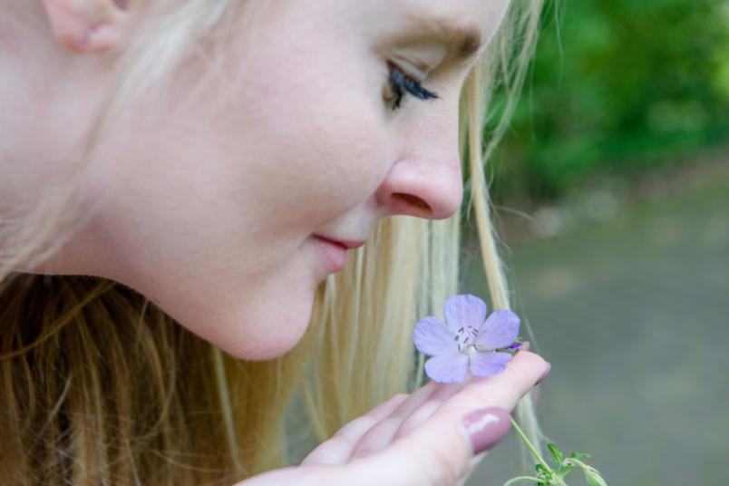 Eine junge Frau mit langen blonden Haaren riecht an einer lilanen Blume.