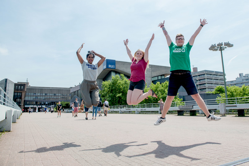 Drei Personen springen mit ausgestreckten Armen, fröhlich auf dem Campus in die Luft.