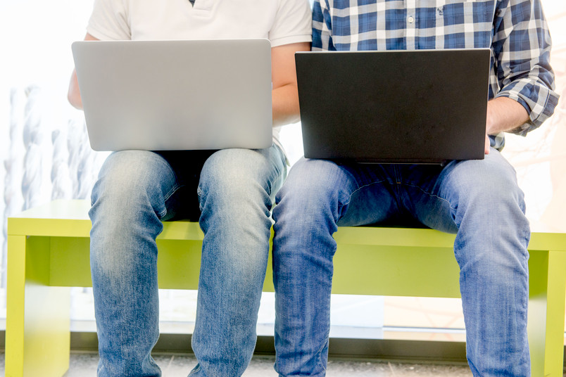 Zwei Männer sitzen nebeneinandern mit Laptops auf dem Schoß. 