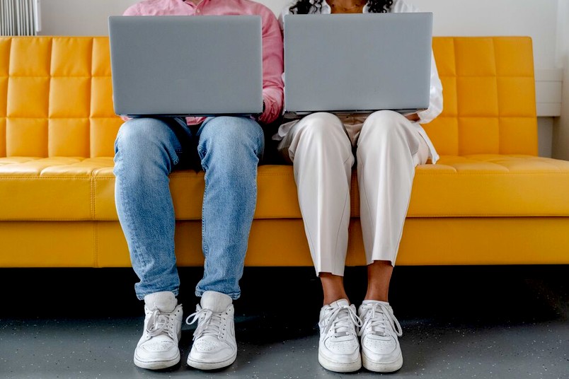 Zwei Studierende sitzen auf einer gelben Couch. Beide haben einen Laptop auf dem Schoß. 