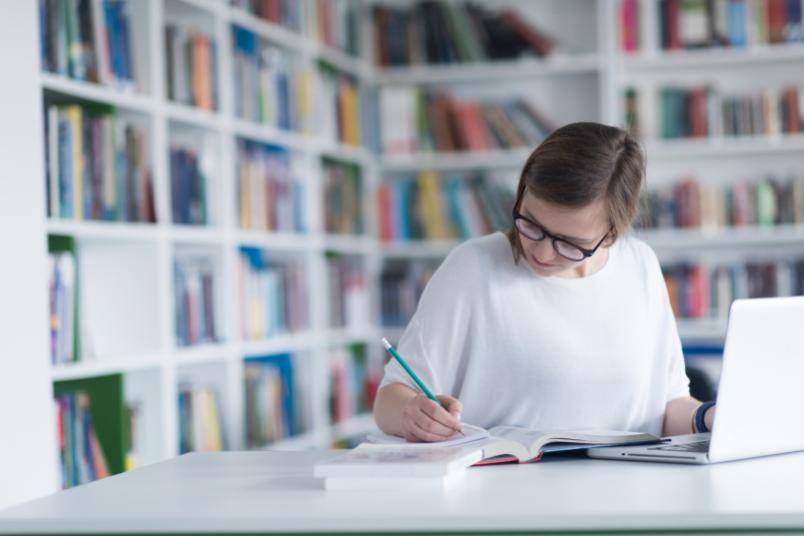 Frau mit Laptop und Büchern macht Notizen in einer Bibliothek