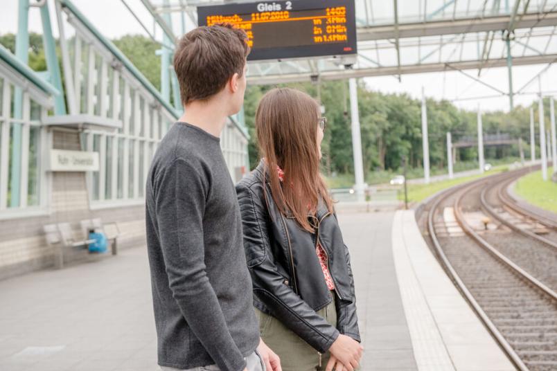 Zwei junge Menschen warten an einer Straßenbahnhaltestelle.