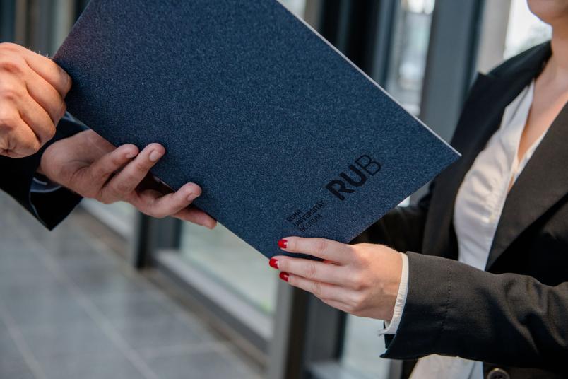 Symbolbild mit einer Urkunde im RUB-Design, die überreicht wird.