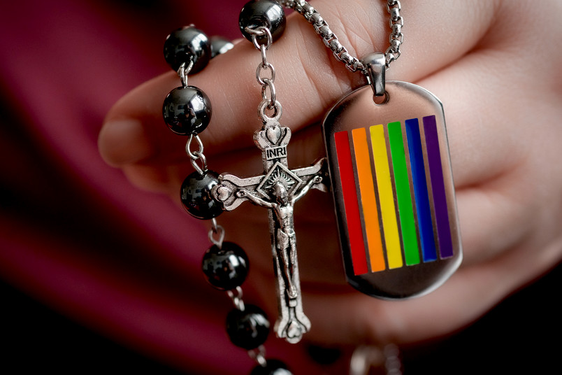 Eine Hand hält einen Rosenkranz, ein Kreuz und einen Anhänger mit Regenbogenflagge.
