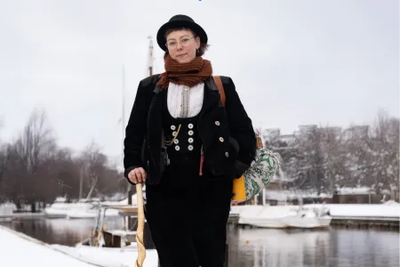 Frau in traditioneller Handwerkerkluft mit Wanderstab vor einem Hafen. 