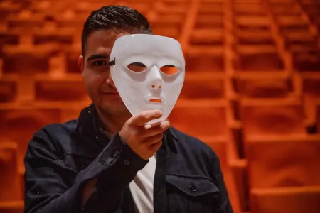 Eine Person hält eine weiße Maske vor das Gesicht, im Hintergrund rote Theatersitze.