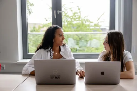 Zwei Frauen sitzt an zwei Laptops nebeneinander und unterhalten sich. 