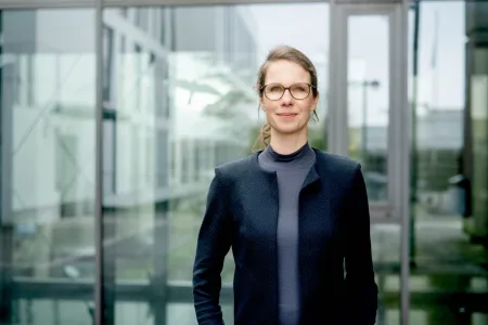 Frau mit dunklem Blazer, Brille und blondem Haar im Zopf