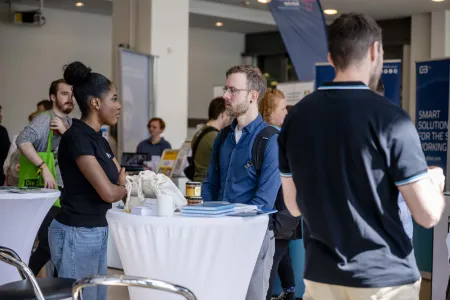 Ein Stehtisch an dem eine junge Frau und ein junger Mann stehen. Im Hintergrund sieht man Messetische und Aufsteller.