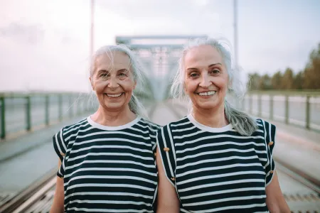Zwei ältere Damen mit weißem Haar und blau-weiß-gestreiften T-shirts stehen nebeneinander. Im Hintergrund sieht man Brückenpfeiler. Die älteren Damen sind ein genetisches Zwillingspaar.