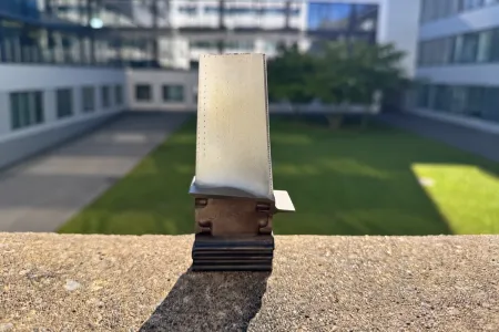 Eine Turbinenschaufe steht auf einer Mauer in der Sonne, im Hintergrund ein grüner Hof