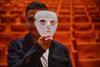 Eine Person hält eine weiße Maske vor das Gesicht, im Hintergrund rote Theatersitze.